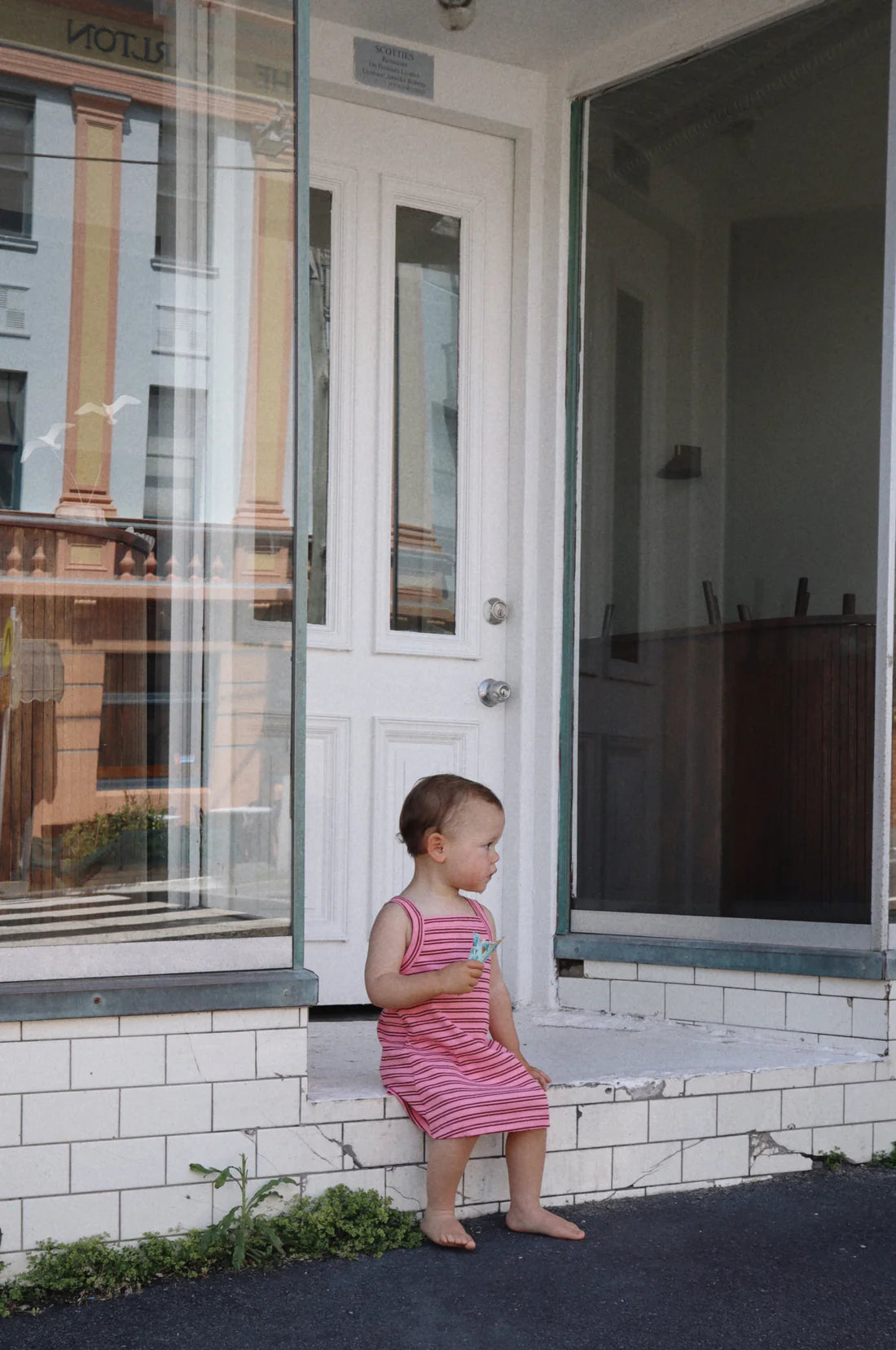 A toddler in a classic MILKY DESIGNS Bobbi Rib Dress in a pink/red stripe pattern sits barefoot on a white tile step in front of a building with large glass windows and a white door. The child gazes off to the side, and the setting resembles the entrance to a shop or café.