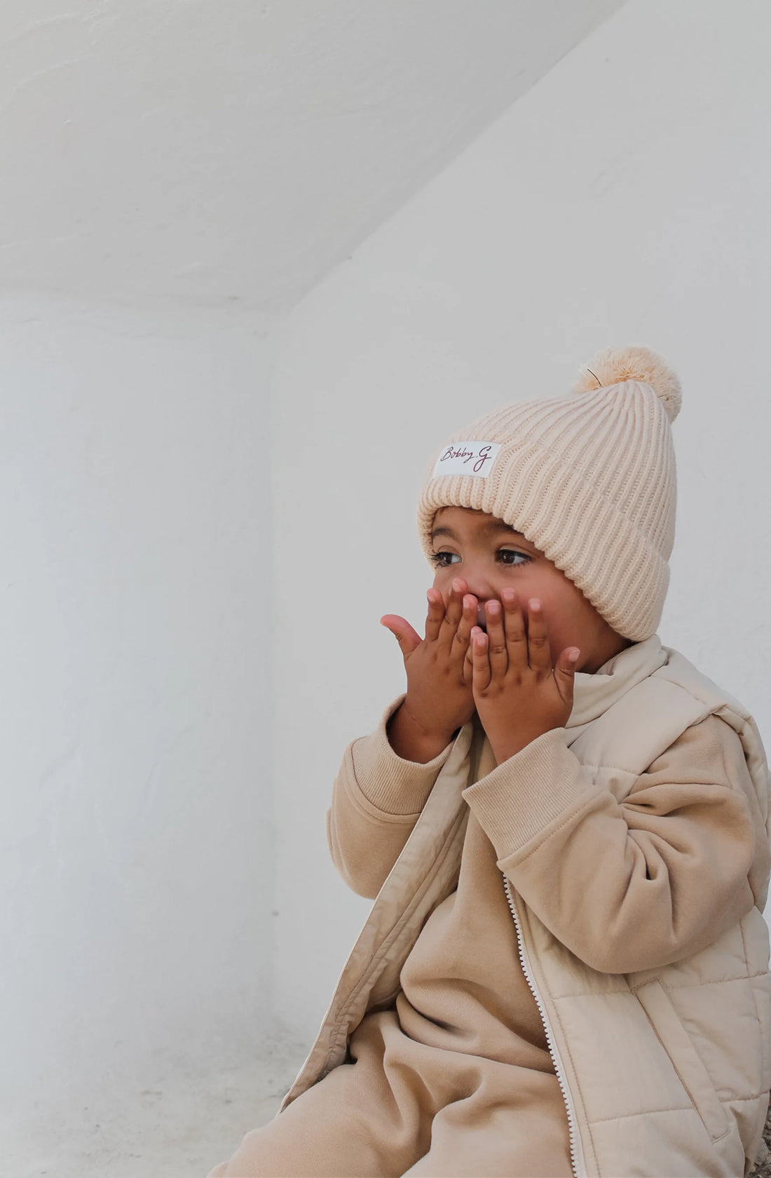 A toddler wearing the ari puffer vest in eggshell with a matching cream jumper and pant set underneath and a cream beanie with the words Bobby G on the front.