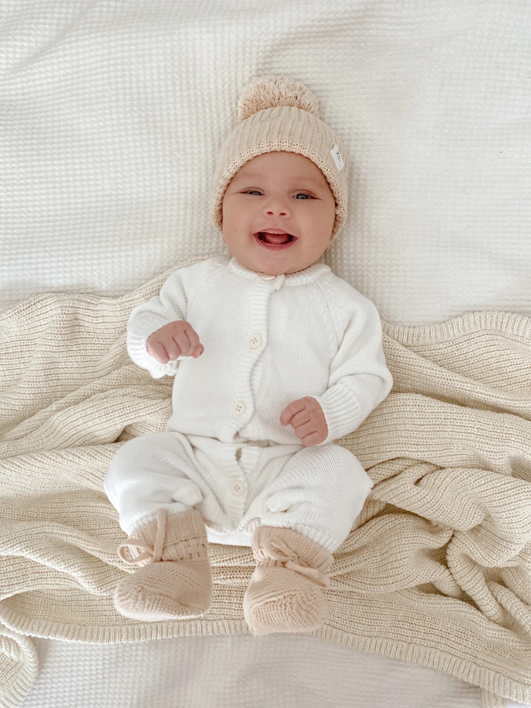 A smiling baby in the Ziggy Lou - Classic Knit Romper Milk by ZIGGY LOU sits on a textured cream blanket, highlighting soft tones—a perfect winter outfit for stylish little ones.