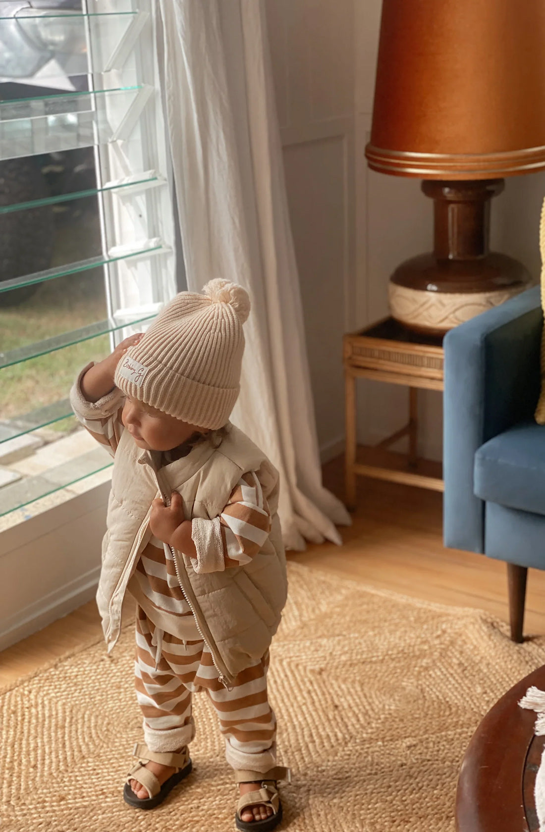 A young toddler wearing the pom pom beanie in creme with a matching brown and white striped jumper and pant set and a puffer vest. The toddler is also wearing sandals and is standing in a room next to a window.