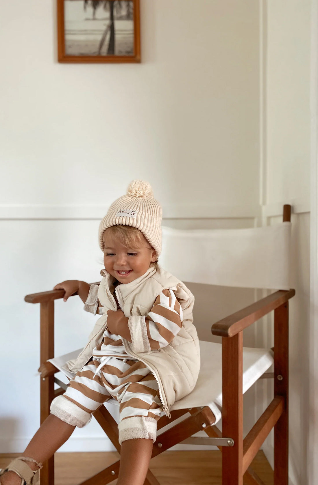 A young toddler wearing the Camden caramel striped pullover and matching pants with a puffer vest over the top and the pom pom beanie in creme. They are sitting on a chair looking down at the floor.