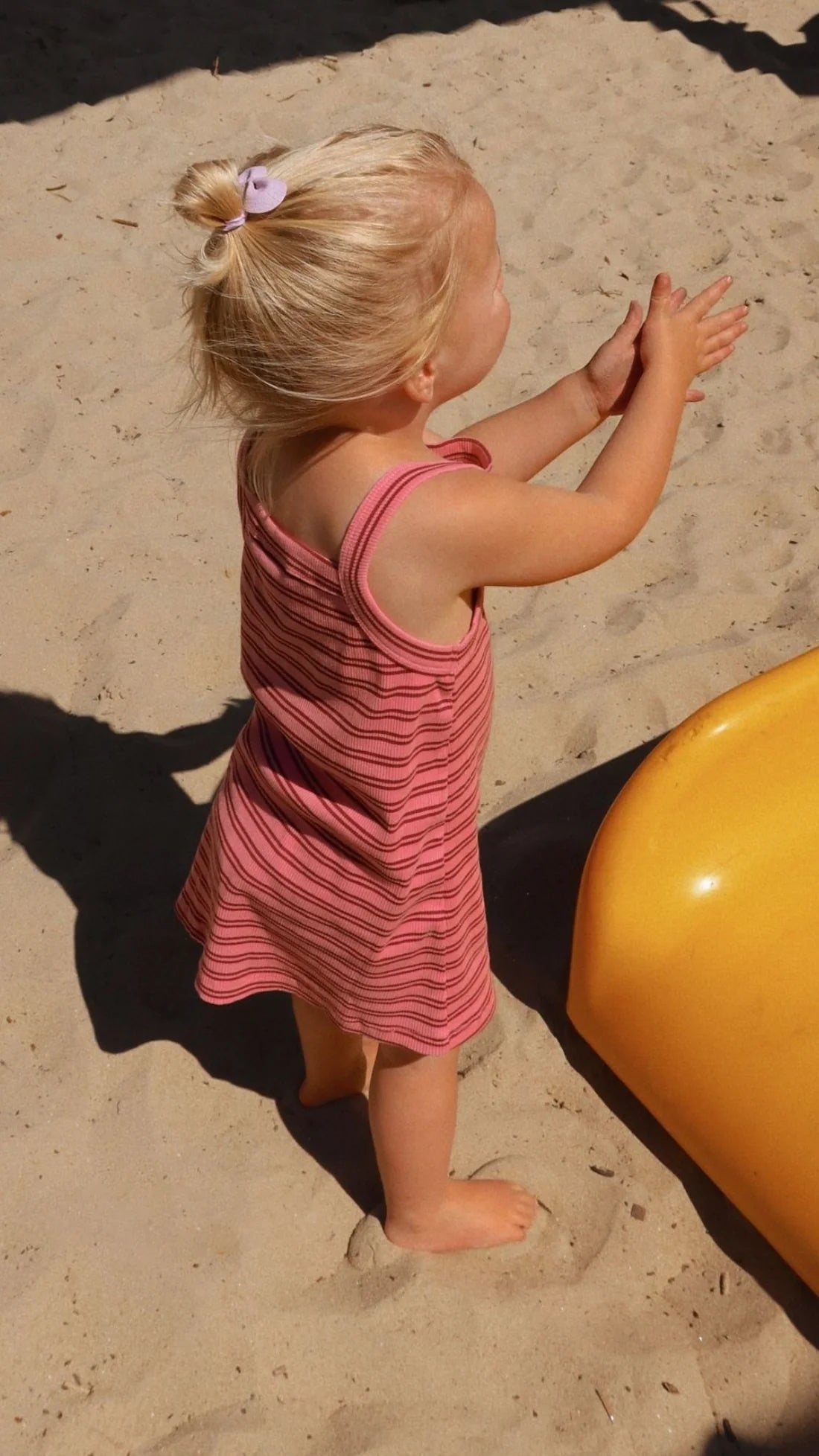 A young child with blonde hair tied into a small bun, wearing a pink and red striped Bobbi Rib Dress by MILKY DESIGNS, stands on sandy ground near a yellow playground slide, clapping hands. The shadows of nearby structures cast patterns on the sand.