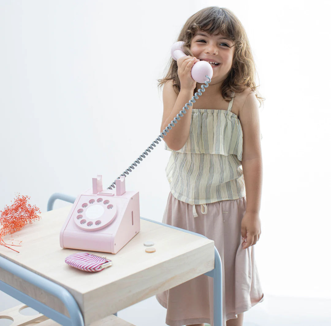 A young girl with long hair is standing by a light-colored table, smiling and holding the KIKO & GG Retro Telephone Pink to her ear. The table is adorned with a small pouch and some decorations. Dressed in a sleeveless striped top and light skirt, she exudes nostalgic retro charm in this bright, minimalistic setting.