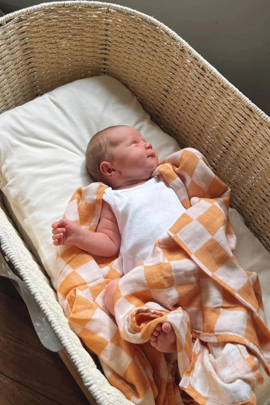 A newborn baby, swaddled in the Apricot Peaches Wrap featuring MILKY DESIGNS' unique textile prints, lies in a woven bassinet. The baby is dressed in a white sleeveless onesie and is looking upwards. The bassinet, lined with a cushion of buttery softness, rests on a wooden floor.