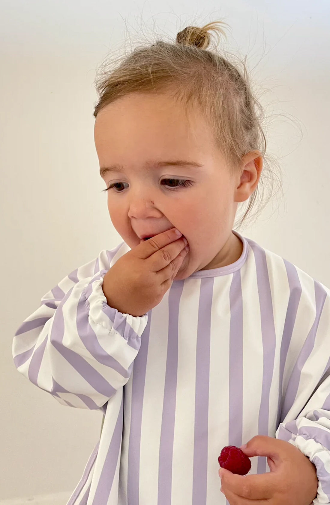 A young girl in the ROMMER Smock Bib in Lilac Stripe eating a rasberry.