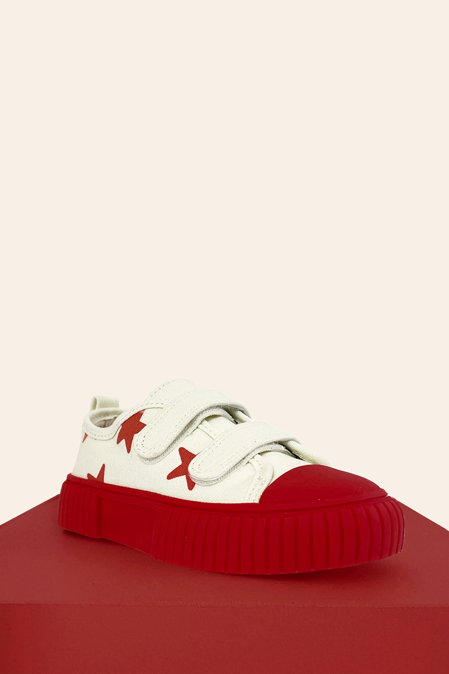 The Limited Edition Piccolini x By Billie Low Top Sneaker in Red Star, from the brand PICCOLINI, is presented on a red platform against a light background. This white sneaker features striking red stars and a matching red toe cap. It includes Velcro straps for easy fastening, a thick non-slip rubber sole for stability, and an organic cotton design that ensures comfort.