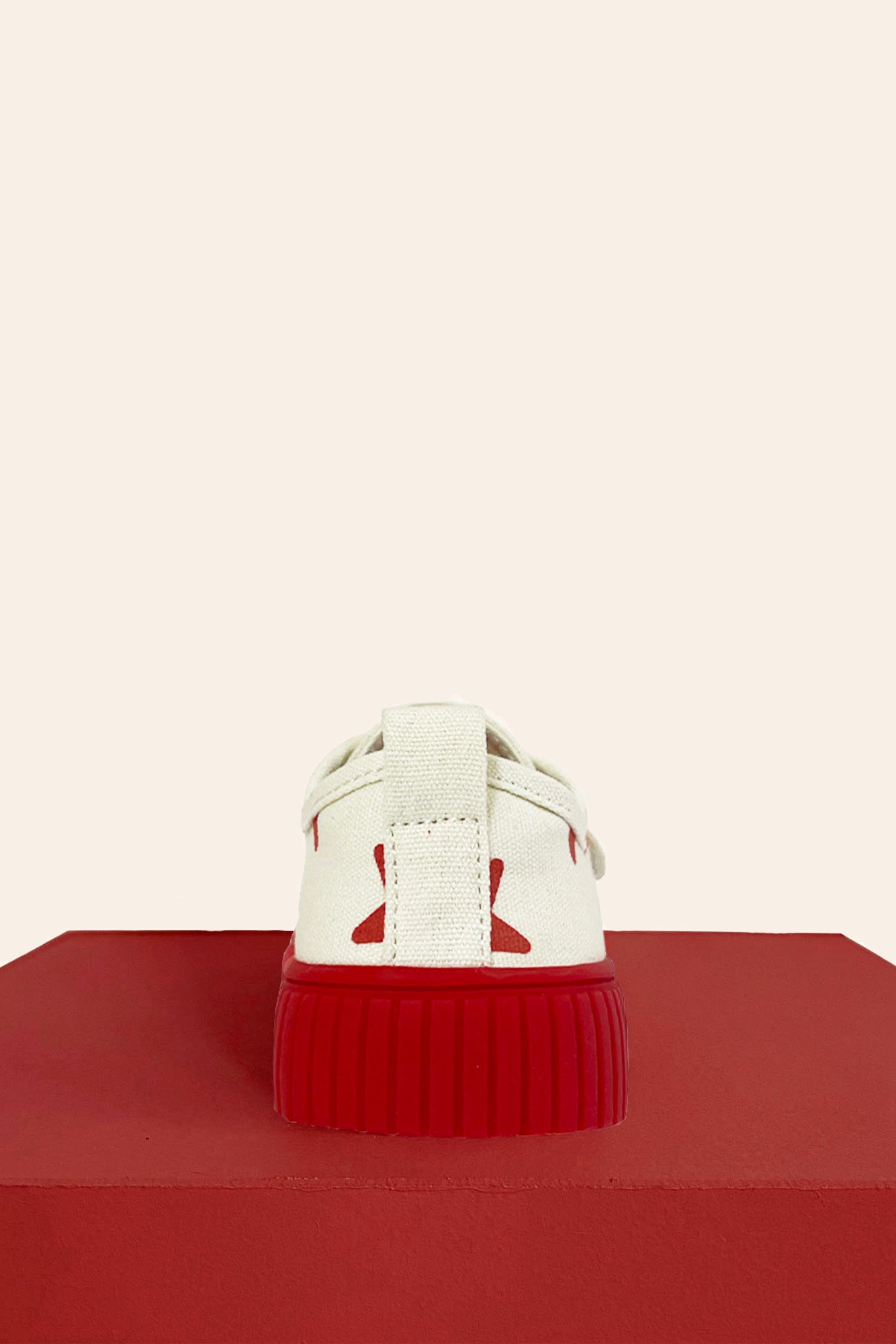 The Limited Edition Piccolini x By Billie Low Top Sneaker Red Star by PICCOLINI, featuring a white design with red accents and a non-slip rubber sole, is showcased from the back on a red platform against a beige background.