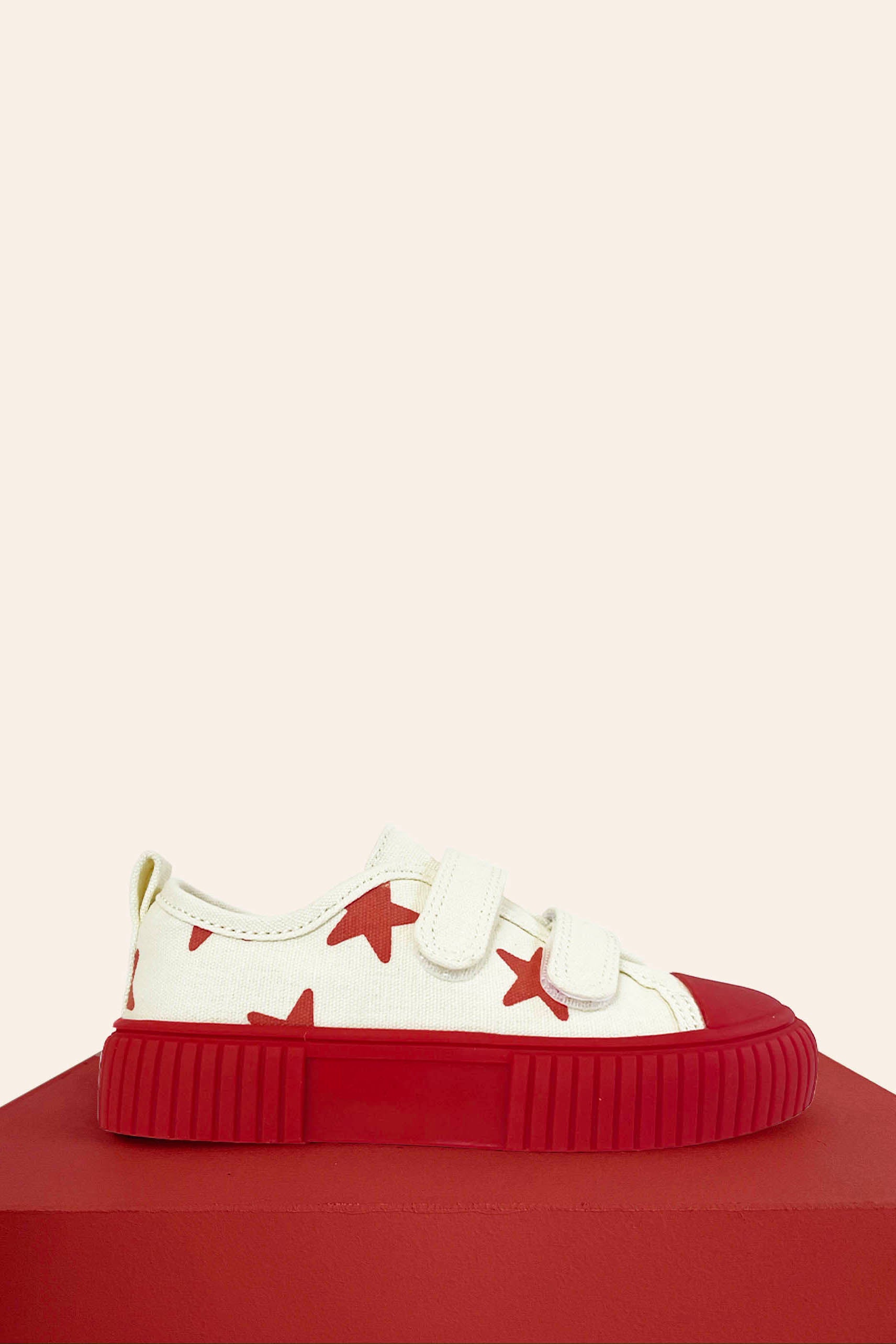 The Limited Edition Piccolini x By Billie Low Top Sneaker Red Star by PICCOLINI is a white sneaker adorned with red stars and equipped with two Velcro straps. It rests on a red platform and features a striking bold red toe cap along with a non-slip rubber sole, all set against a neutral background.