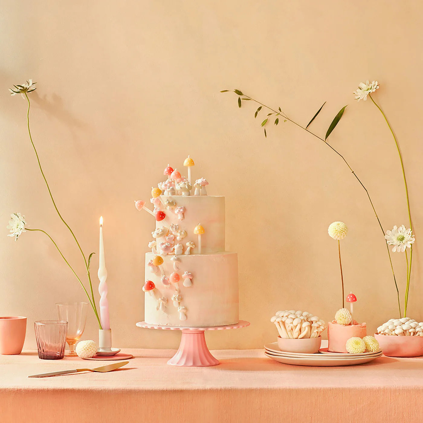 A beautifully decorated cake, featuring pastel flowers and mushrooms, takes center stage on a stand, accompanied by MERI MERI Mushroom Birthday Candles from a pack of six in two sizes. Surrounded by elegant tableware and floral arrangements, it creates the perfect setting against a soft peach-colored wall for an enchanting autumnal party.