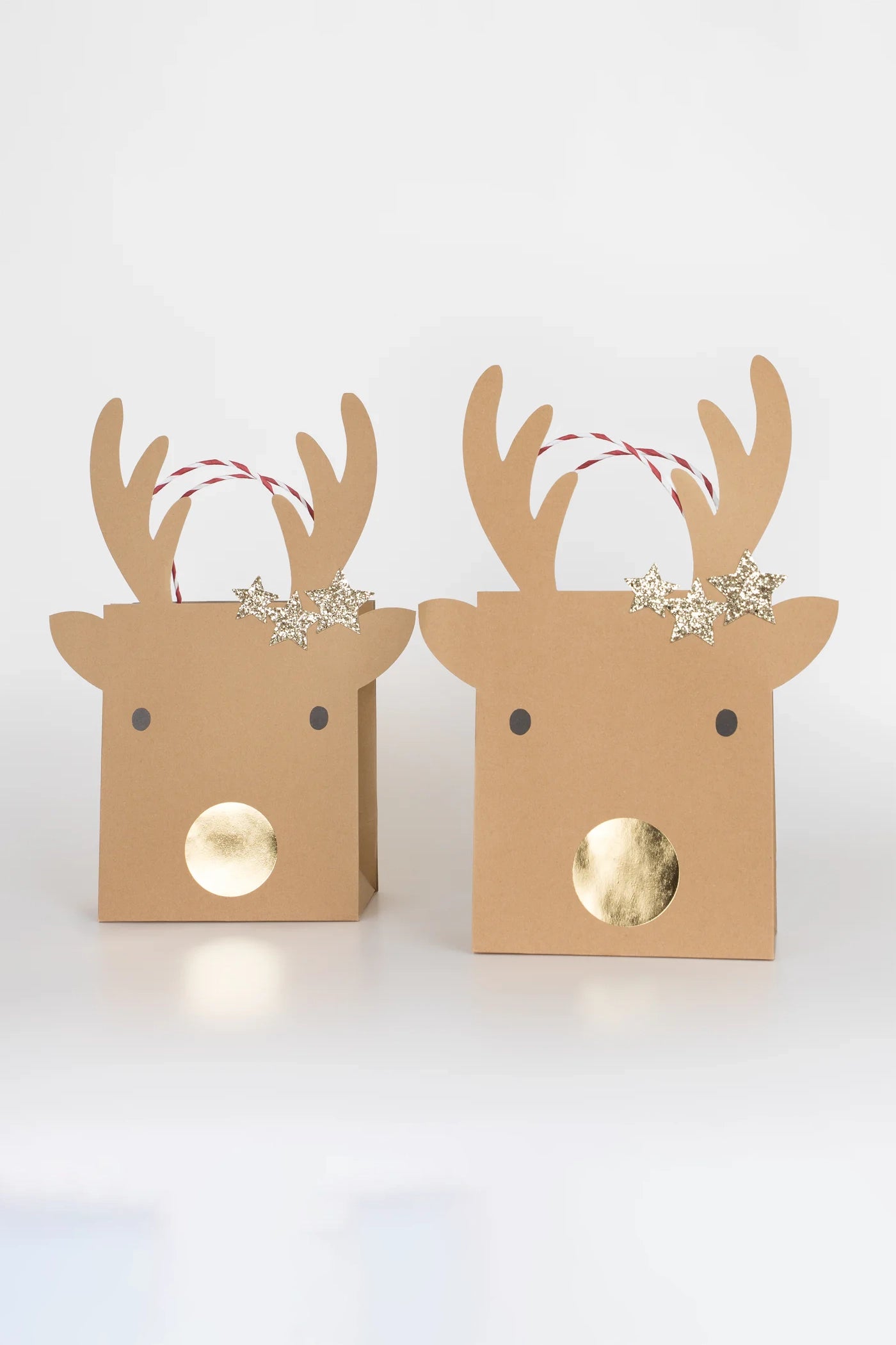 Pack of two medium-sized gift bags from MERI MERI, featuring a brown reindeer face design with antlers and red-and-white striped handles. The bags are adorned with gold foil noses and decorative stars near the antlers against a plain white background.