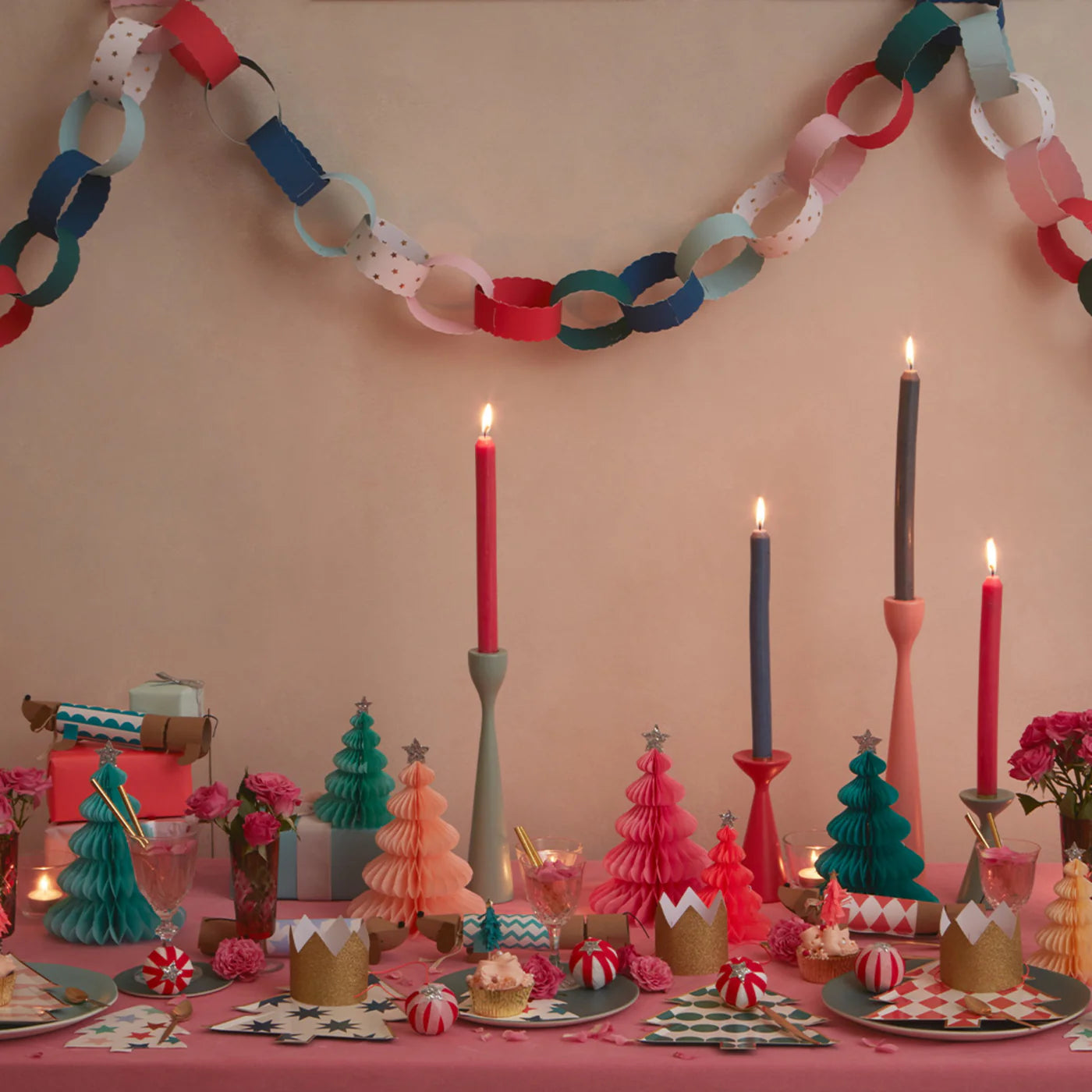 A festive table adorned with MERI MERI's Rainbow Forest Honeycomb Decorations, complete with colorful trees and enhanced by the glow of candles, pink flowers, paper crowns, and candy. Above, a garland of multicolored paper chains sways gracefully. Plant-based glitter adds a touch of sparkle to these vibrant and playful tissue paper decorations.