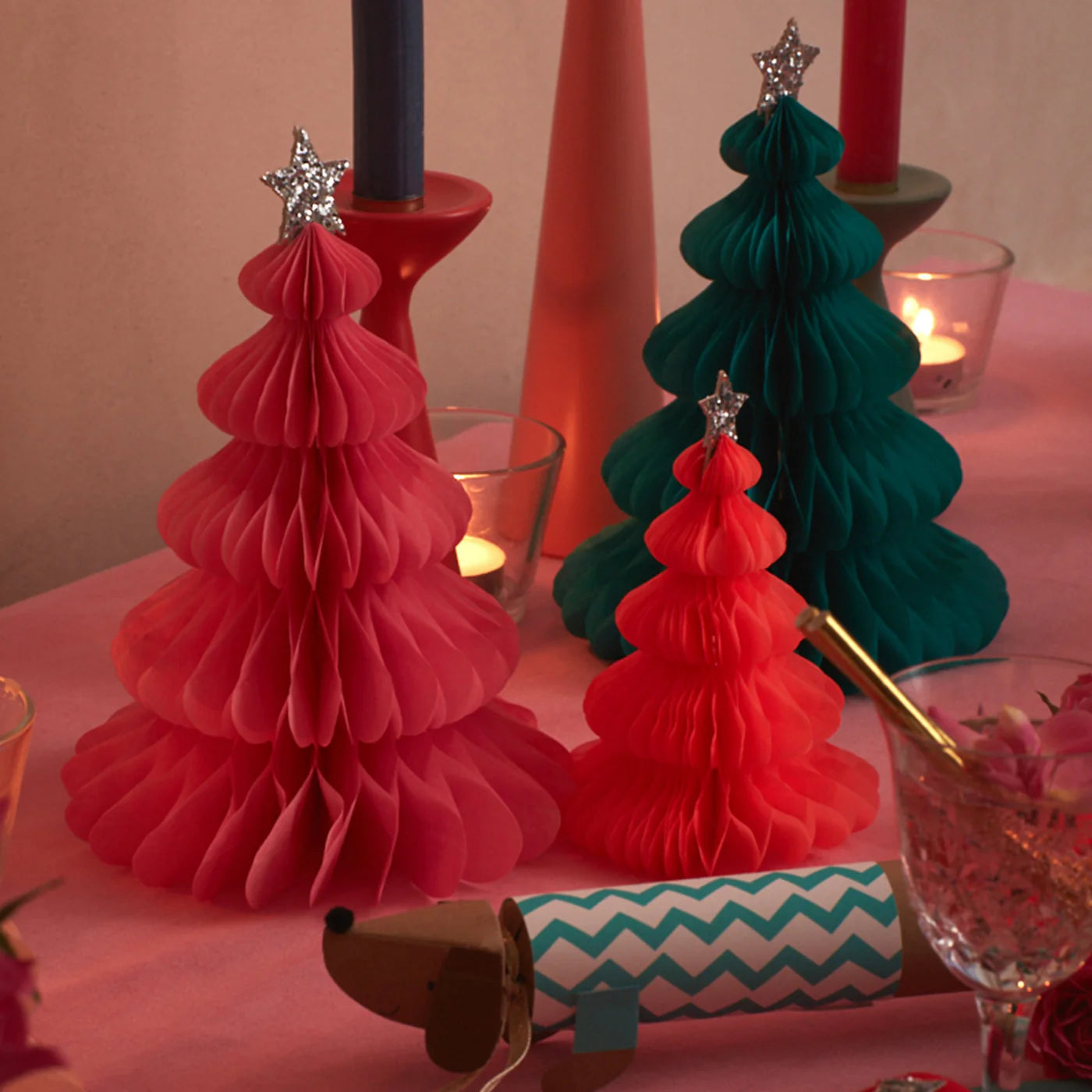 A festive holiday table setting features the vibrant MERI MERI Rainbow Forest Honeycomb Decorations, showcasing an array of colors with star toppers. These tissue paper decorations enhance the cheerful scene, while candles and a party cracker complete the cozy ambiance.