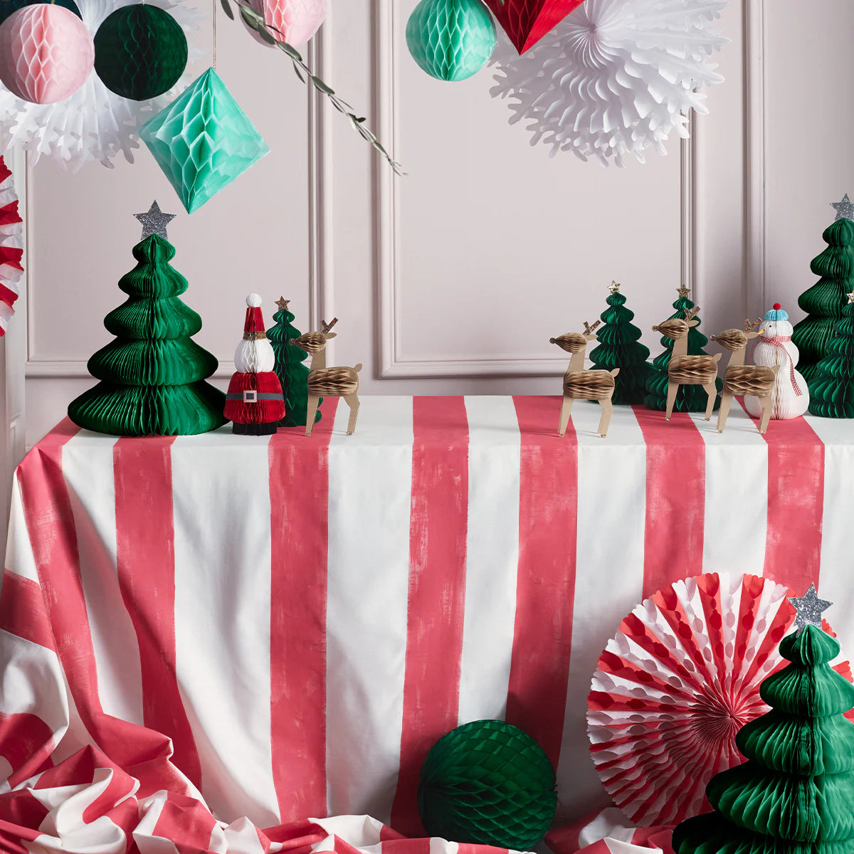 A festive display features a table covered with a red and white striped cloth, adorned with green Giant Honeycomb Trees from MERI MERI. Reindeer figurines and Santa decorations enhance the scene while glittery stars hang among the ornaments, adding holiday charm to a classic backdrop.