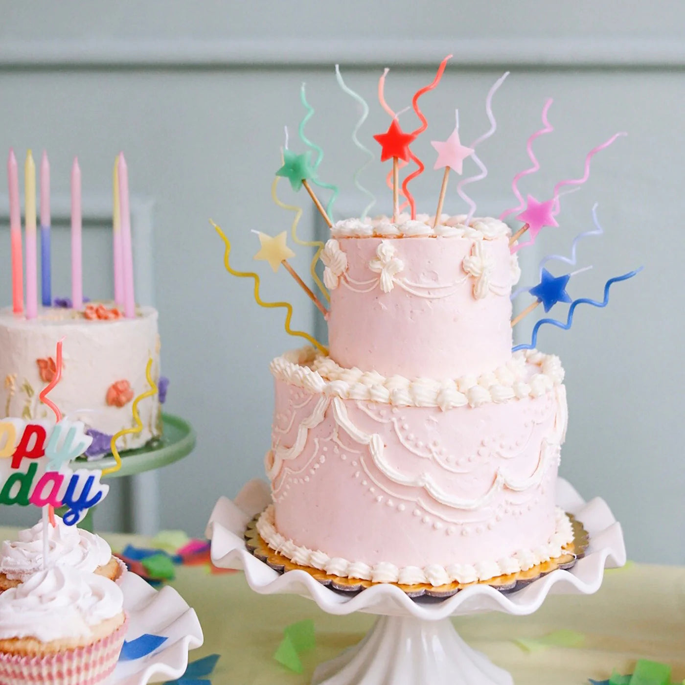 A pink two-tiered cake with intricate frosting details and colorful star-shaped toppers sits elegantly on a white stand. In the background, a celebration cake shines with vibrant MERI MERI Pastel Swirly Candles, available in a pack of 20 in six colors, complemented by cheerful cupcakes. The atmosphere is festive and lively.