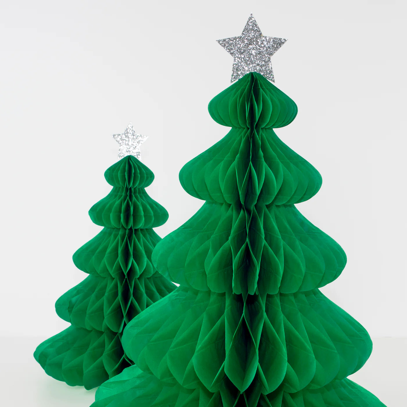 In front of a plain white background, two green honeycomb Christmas trees from the MERI MERI Giant Honeycomb Trees pack are displayed, each adorned with a glittery silver star. The trees come in varying sizes, with one larger tree prominently positioned in the foreground.