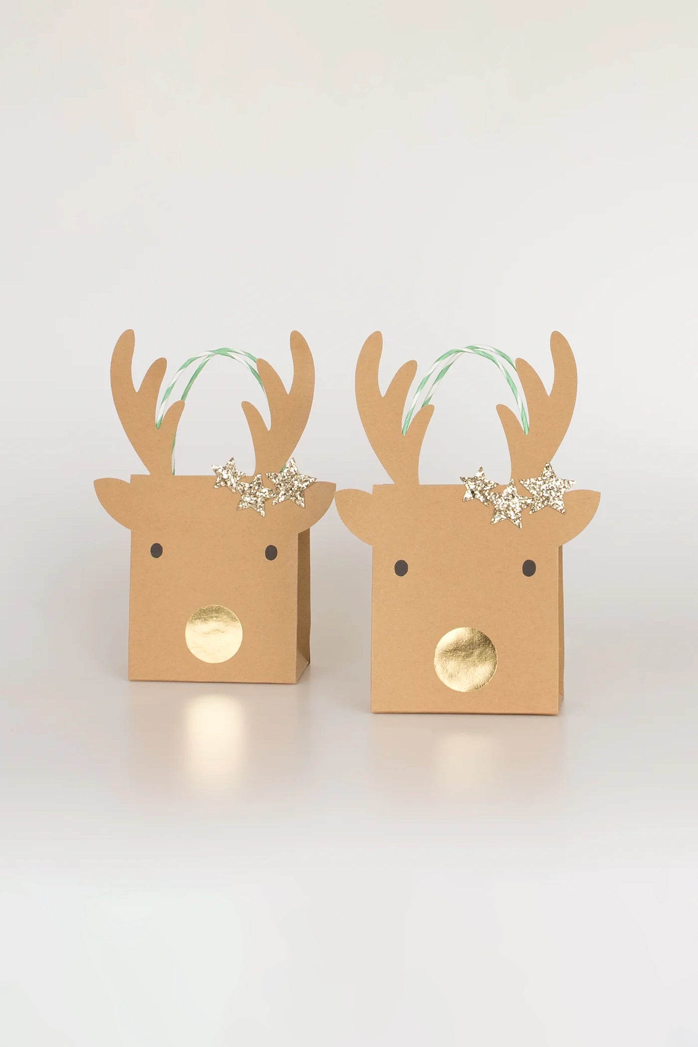 Two Small Reindeer With Stars Gift Bags by MERI MERI, styled to resemble reindeer faces with charming antlers and gold noses. Each bag is adorned with gold glitter stars on the head and comes with green-and-white striped handles, making them a delightful addition for holiday cheer against a plain white background.