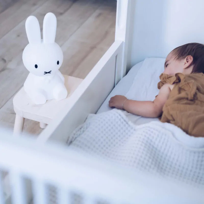 A young child sleeps peacefully in a crib, covered by a white blanket. Near the crib, on a small wooden stool, sits an MR MARIA Miffy First Light Lamp with black eyes and a simple expression. The background features a light wooden floor.