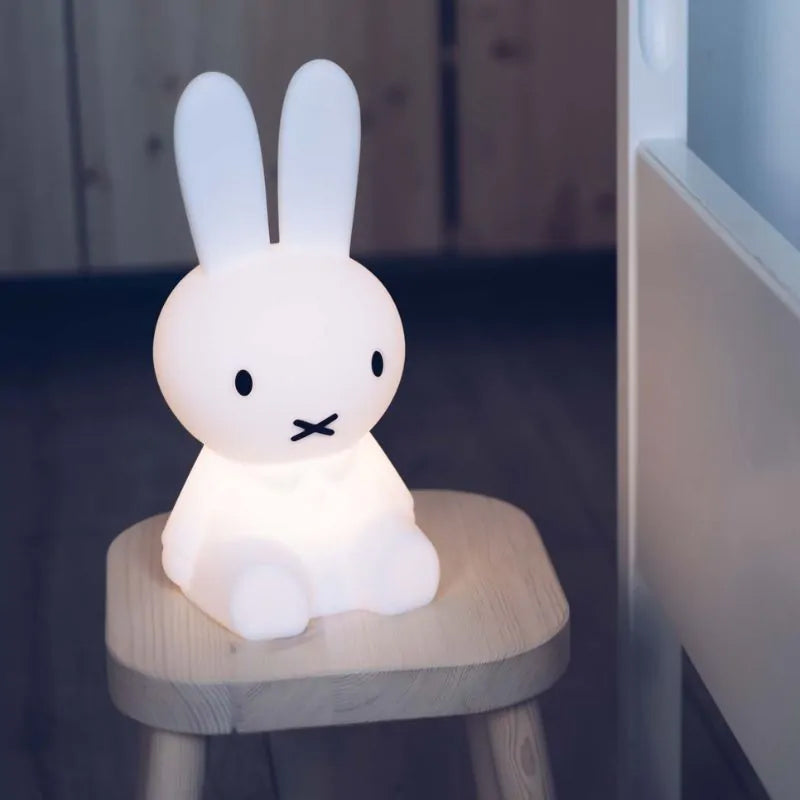 The MR MARIA Miffy First Light Lamp, shaped like a white rabbit, rests on a wooden stool in a softly lit room. The rechargeable LED emits a gentle, warm glow that illuminates the surrounding area. The background includes child-safe wooden furniture.