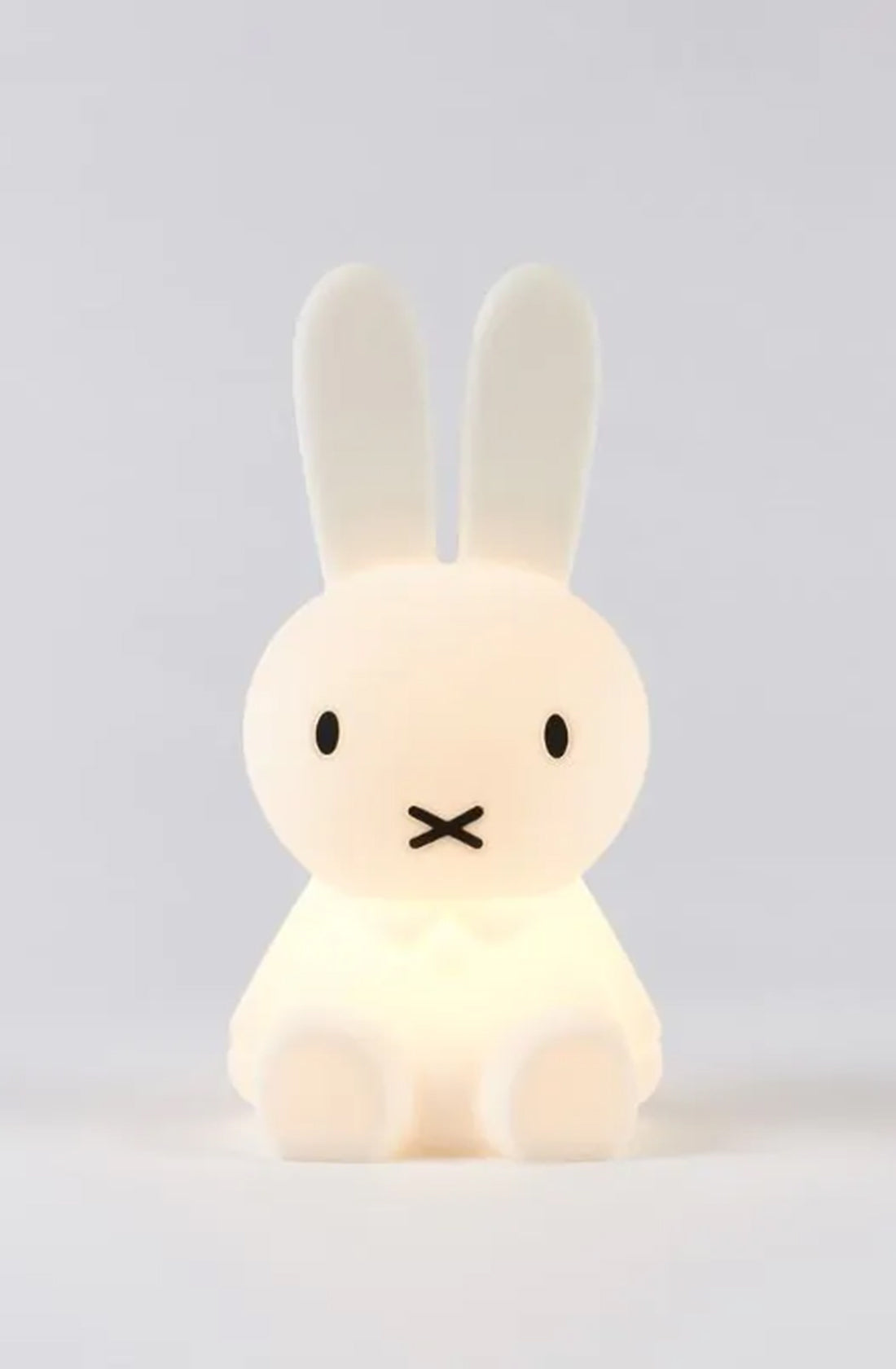 The MR MARIA Miffy First Light Lamp is a small, white night light shaped like a sitting cartoon bunny with long, upright ears and black, simple facial features, featuring two dot eyes and an “X” for a mouth. Set against a light gray background, this rechargeable LED lamp is child-safe and perfect for any nursery.