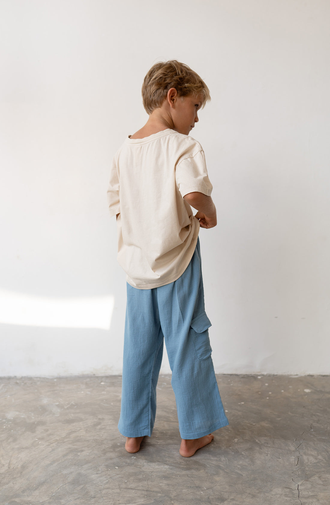 Young boy wearing the henna pants in cornflower and a beige t shirt.