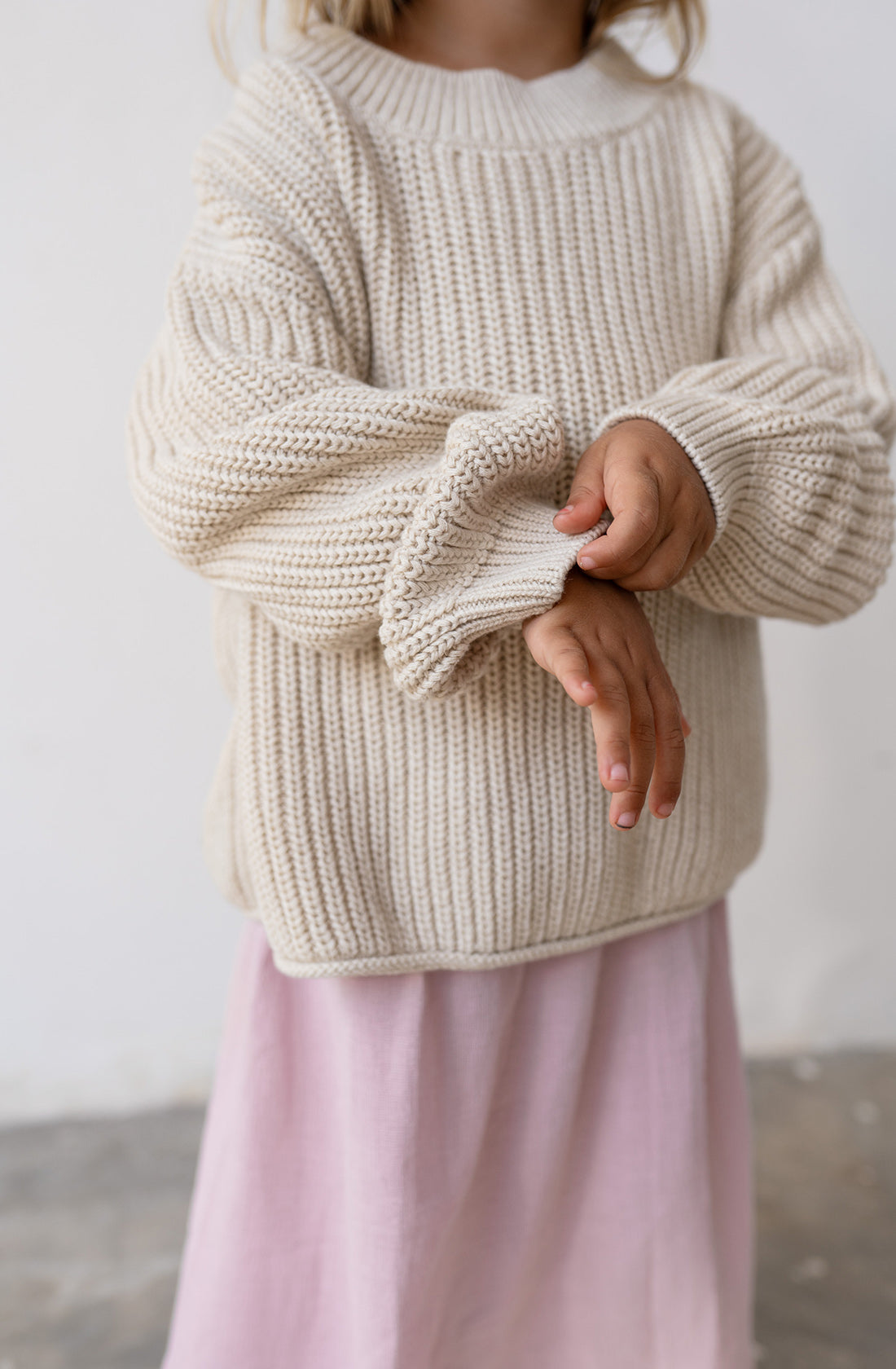 A young girl folding the sleeve of the chunky knit jumper in natural.