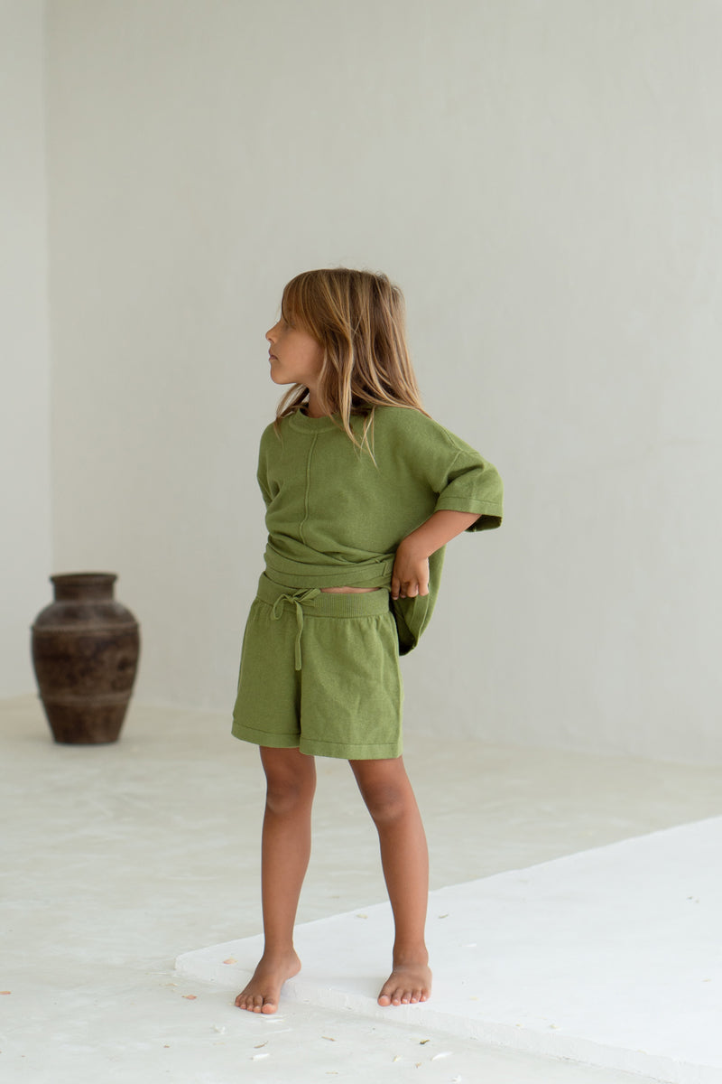Girl wearing knit tee and short set in olive, showing the short waistband by lifting up the tee 