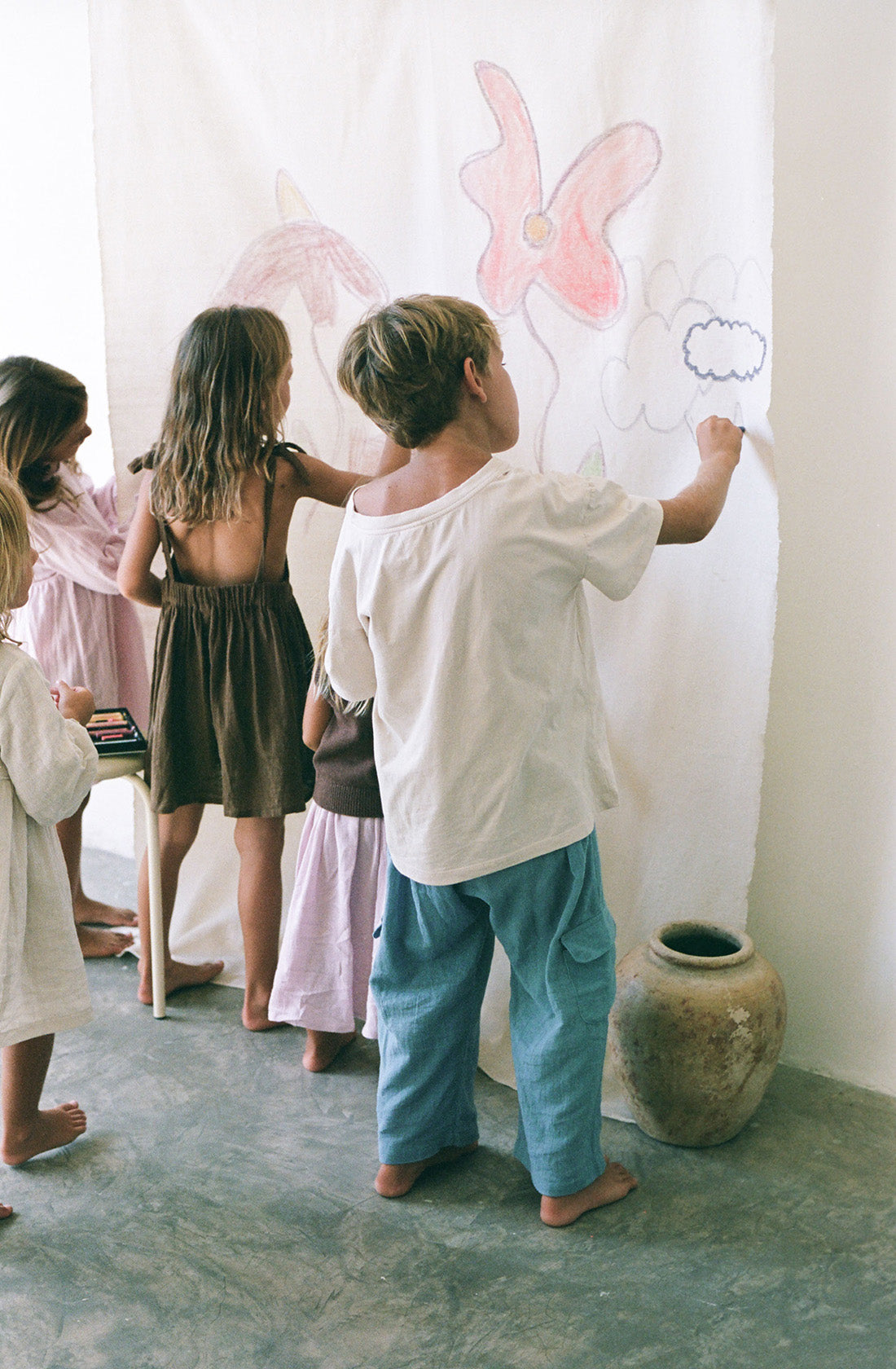 Young boy wearing the henna pants in cornflower and a beige t shirt while drawing on a canvas.