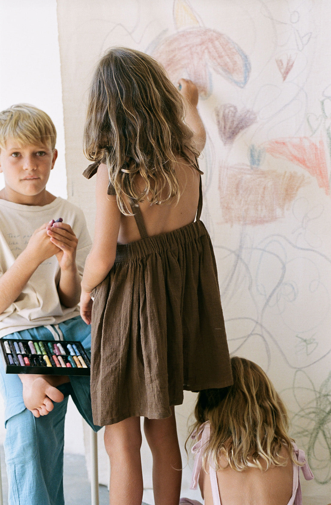 Young girl wearing the luna pinafore in cocoa while drawing on a canvas with some crayons.