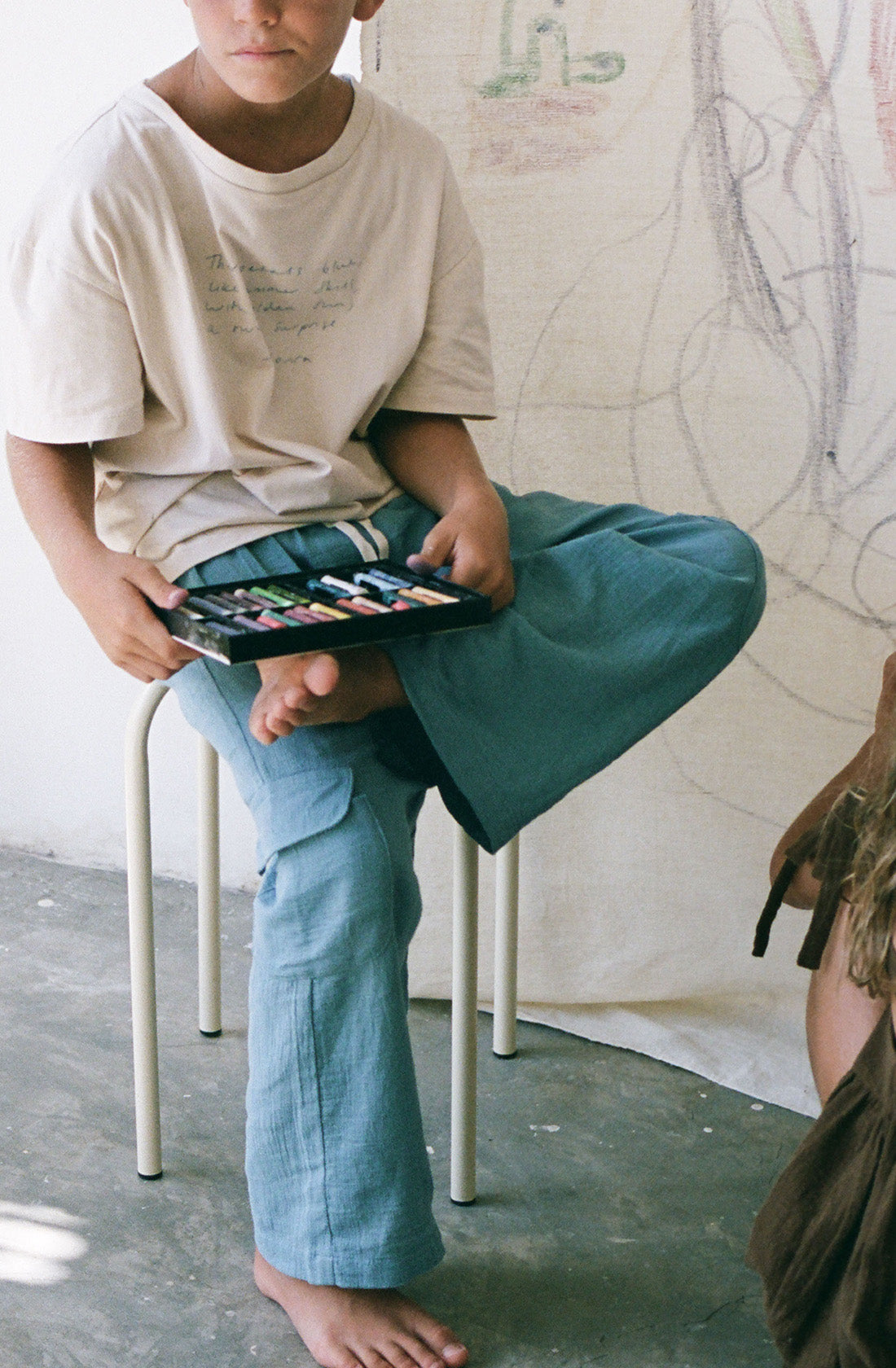 Young boy wearing the poet tee undyed and the henna cornflower pants holding a set of crayons.