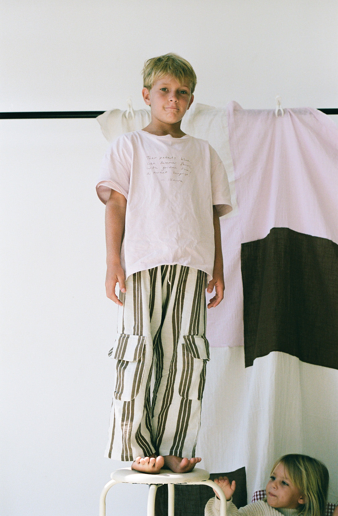 Young boy wearing the henna pants in cocoa stripe with a pink shirt standing on a chair.