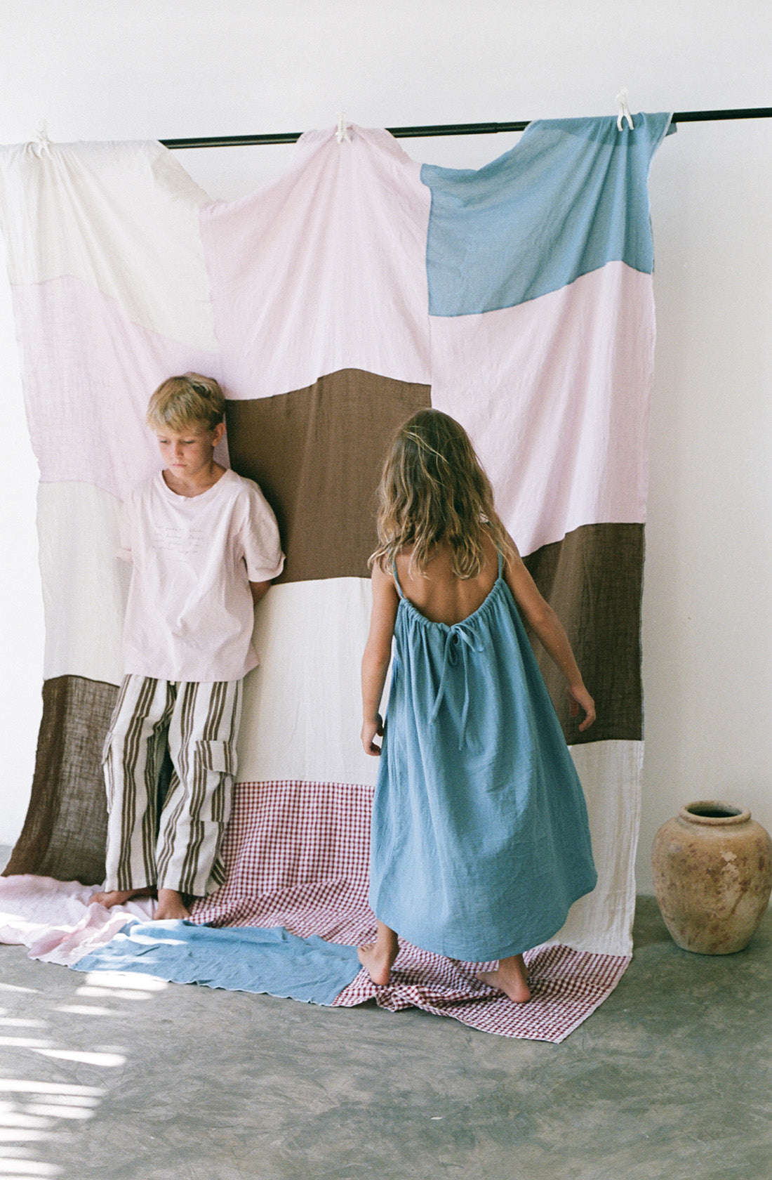 Young boy wearing the henna pants in cocoa stripe with a pink t shirt standing in front of a checkered blanket.