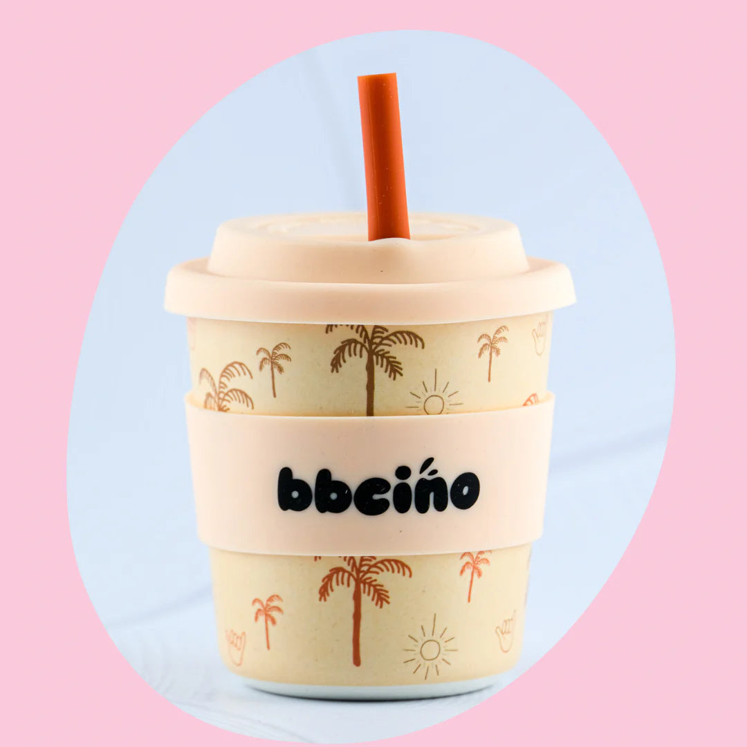 The Reusable BambinoCino Cup 240ml Oasis by BBCINO is a beige cup decorated with palm tree and sun designs. It includes a lid, reusable silicone straw, and a silicone grip sleeve with "bbeño" written on it. The background features a light pink oval shape and a blue area peeking out on the right side.