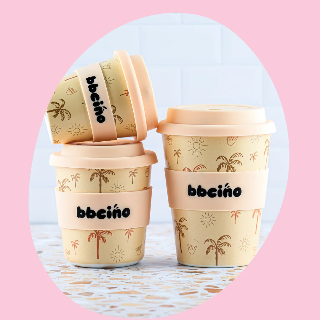 A set of Beige Reusable BambinoCino Cups (240ml Oasis) with matching lids and palm tree and sun illustrations. The cups feature the word "BBCINO" in black on the front, alongside a reusable silicone straw, all set against a pink oval background.