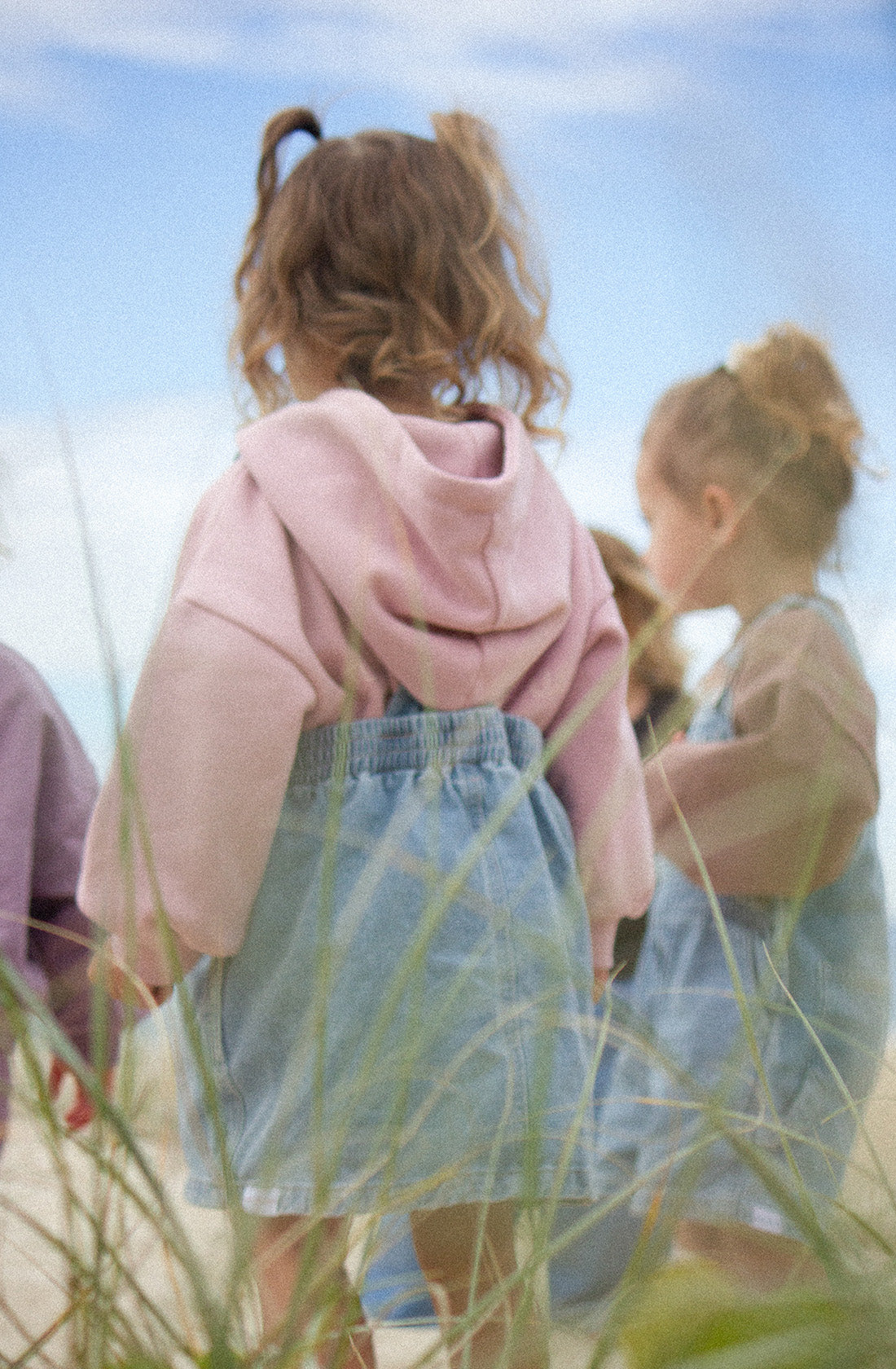 A group of young children is standing outdoors on a slightly cloudy day. The focus is on a child in a pink Logo Hoodie Set Pink Haze by TWIN COLLECTIVE and denim overalls, facing away from the camera. Other children are visible in the background, also wearing casual, comfortable clothing.