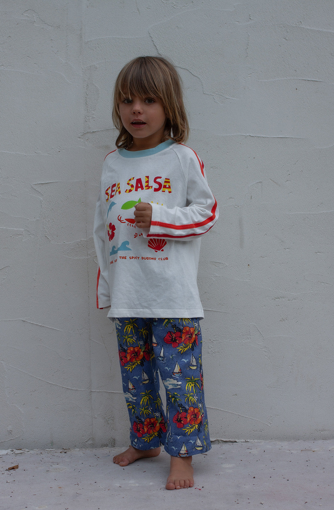 A young child with long hair stands barefoot against a plain wall. The child is wearing a 100% cotton, SPICY DUGONG Sea Salsa Long Sleeve Tee featuring a fun sea-themed print and blue pants adorned with a tropical design. The child appears to be holding a small green object.