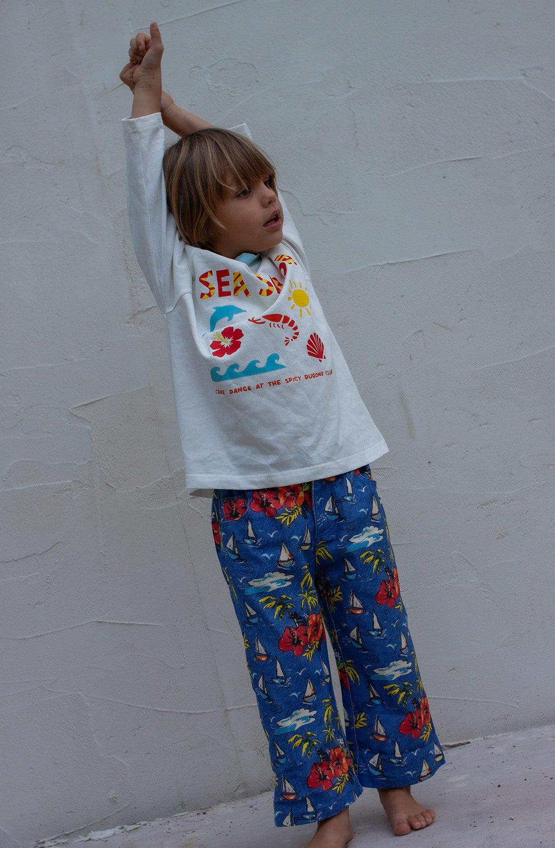 A young child with blonde hair is stretching in a SPICY DUGONG Sea Salsa Long Sleeve Tee, showcasing colorful graphics of the ocean, sun, and the text "SEA DAY." They are also wearing blue pants adorned with vibrant red flowers and sailboats, standing against a plain, light-colored wall.