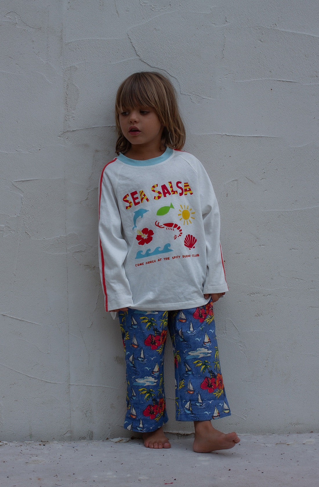 A young child with long hair stands barefoot against a plain gray wall. The child is wearing the SPICY DUGONG Sea Salsa Long Sleeve Tee made from 100% cotton, featuring a fun Sea Salsa print, along with blue pants adorned with sailboats and floral patterns. The child looks slightly to the side, showcasing the playful design.