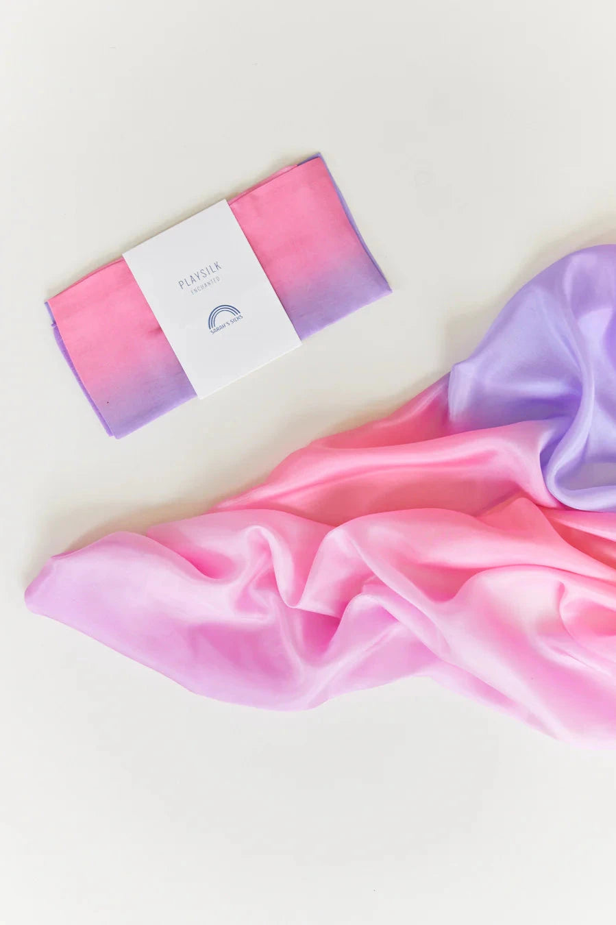 A silk scarf with a pink and purple gradient, similar to the delightful Enchanted Playsilk Blossom by SARAH'S SILKS, is gracefully draped on a white surface. A neatly folded section is packaged with a white label showcasing blue text and the brand's logo, sparking the imagination.