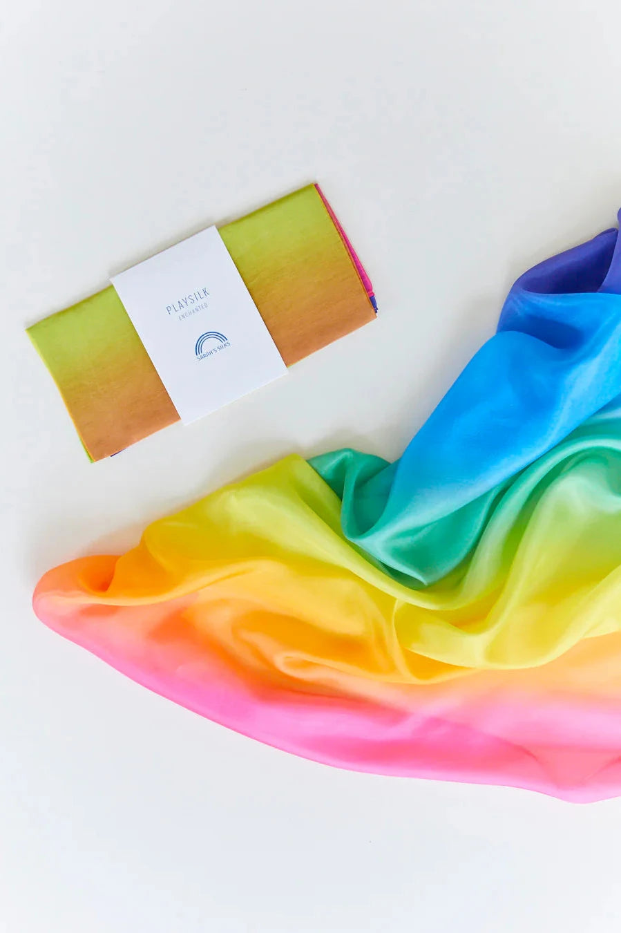 The Enchanted Playsilk Rainbow by SARAH'S SILKS is gracefully showcased on a white background. Made from 100% pure silk, this scarf seamlessly blends shades of red, orange, yellow, green, blue, and purple. Designed for open-ended play, it features a folded section fastened with a white paper band.