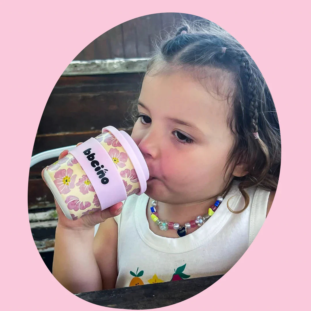 A young child with braided hair drinks from a BBCINO Reusable BambinoCino Cup 240ml Poppy, using a reusable silicone straw. The child is wearing a colorful beaded necklace and a white sleeveless top. The image has a pink oval border.