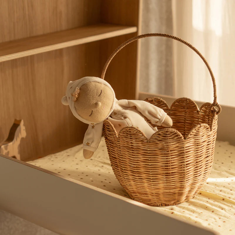 A soft, plush doll dressed in a light-colored hooded onesie lies in an OLLI ELLA Rattan Tulip Carry Basket Natural with a scalloped edge, placed on a dotted crib sheet. A wooden horse toy stands in the background, and warm lighting gives the nursery an heirloom-quality cozy feel.