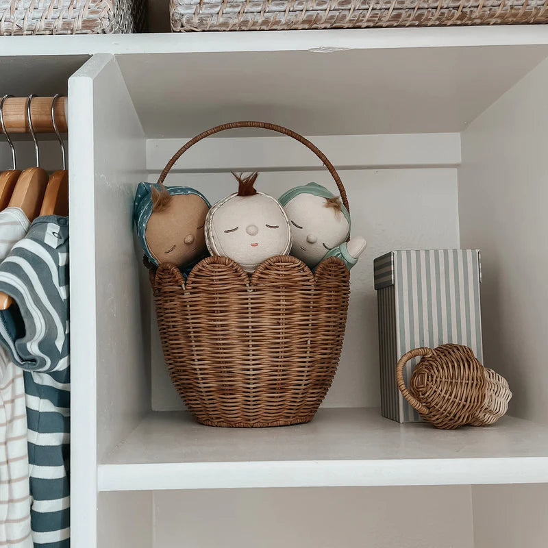 An OLLI ELLA Rattan Tulip Carry Basket Natural, handwoven with natural rattan and filled with three small plush dolls, sits on a white shelf in a closet. To the right of the basket, there is a striped storage box and a smaller wicker basket. Hanging striped clothing is partially visible on the left side of the image.