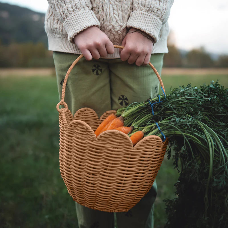 A person wearing a cream sweater and green pants is holding an OLLI ELLA Rattan Tulip Carry Basket Natural filled with freshly picked carrots. The person is standing outdoors in a field with greenery and hills in the background.