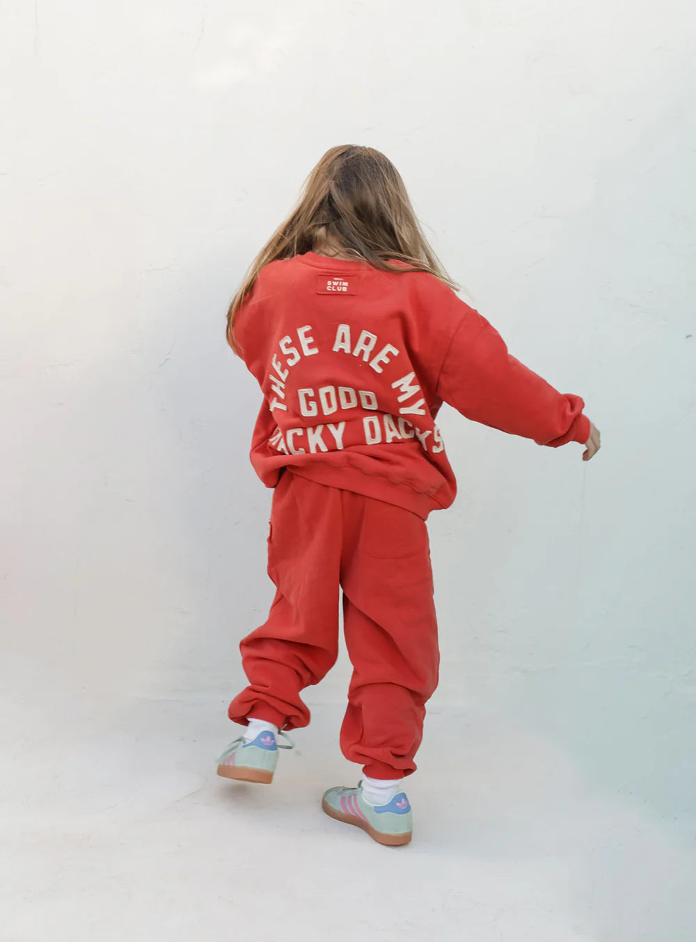 A child in oversized, fleece-lined Small Swim Club - Good Tracky Dacks Red/Cream by SMALL SWIM CLUB stands facing a white wall, arms slightly outstretched, wearing a red sweatshirt with "THESE ARE MY GOOD LUCKY DAYS" on the back and colorful sneakers.