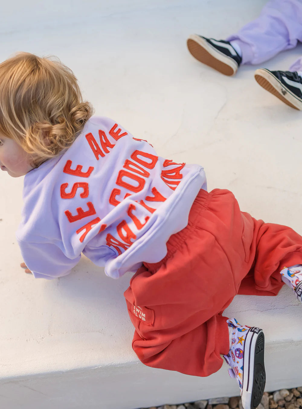 A young child with light brown hair crawls on a white surface, wearing a lavender shirt and SMALL SWIM CLUB Good Tracky Dacks Red/Cream pants. Another child in lavender pants and black sneakers sits nearby.