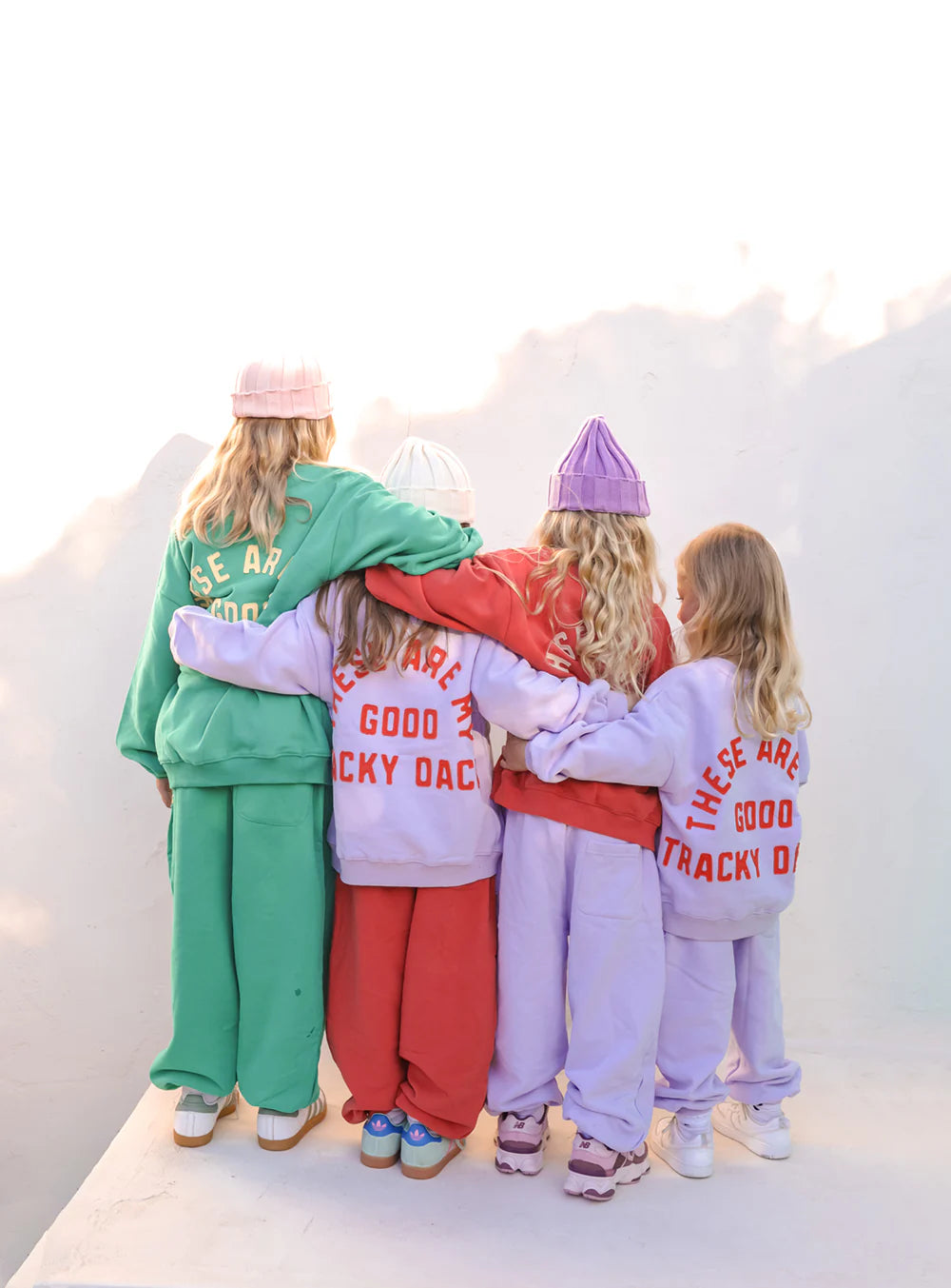 Four children, arms around each other, face away from the camera wearing colorful SMALL SWIM CLUB Good Tracky Dacks Red/Cream sweatpants and fleece-lined beanies in green, red, purple, and pink.