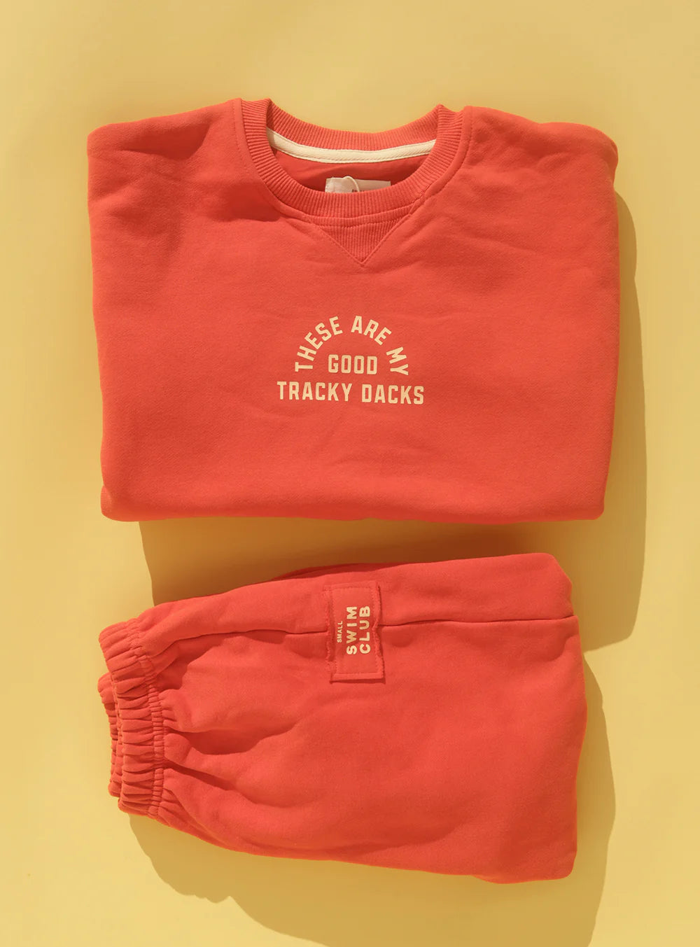 The SMALL SWIM CLUB Good Tracky Dacks Red/Cream set features a folded coral fleece-lined sweatshirt reading "These are my good tracky dacks" and matching oversized slouchy pants with a "Good dacks only" patch, displayed on a yellow background.