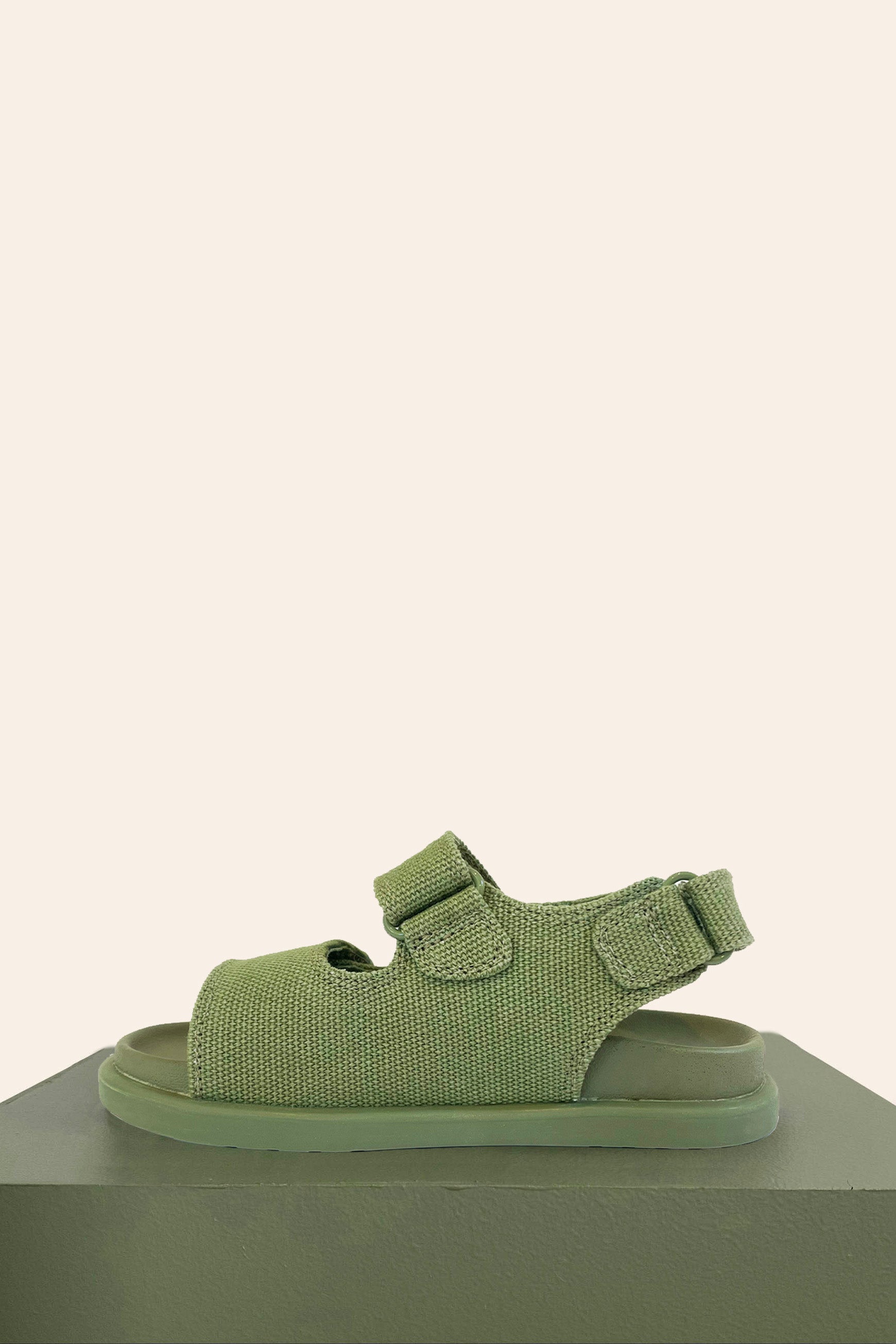 The PRE-ORDER Original Sandals Khaki by PICCOLINI, featuring a textured fabric upper crafted from organic cotton and two adjustable straps, is showcased on a green surface against a beige background. This sandal offers an open toe design and includes a podiatrist-recommended non-slip rubber sole for enhanced stability.