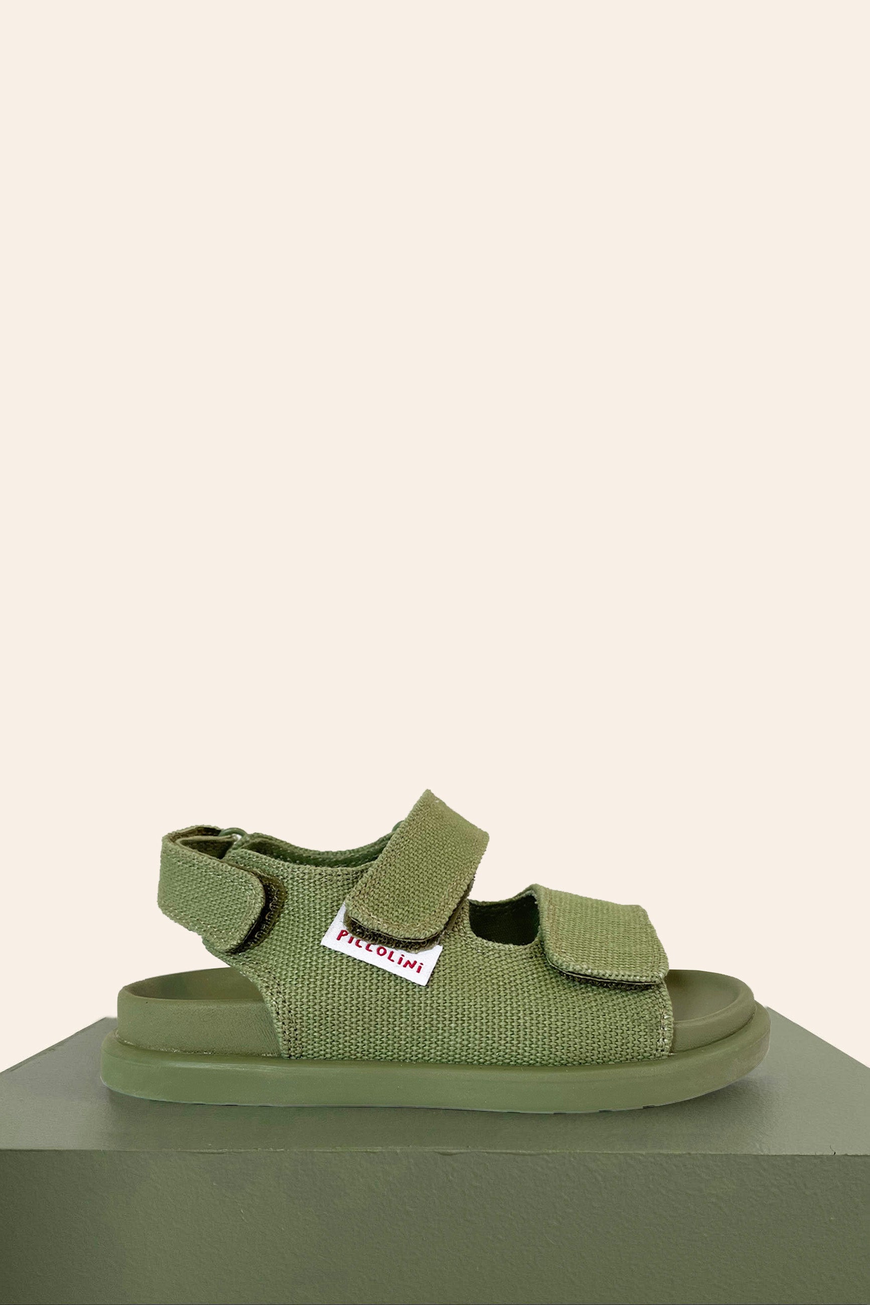 A single khaki sandal from PICCOLINI's PRE-ORDER Original line is showcased against a beige background. It features a textured fabric upper crafted from organic cotton, complete with two Velcro straps. The sandal has a thick, cushioned sole and a small logo tag on the upper strap.