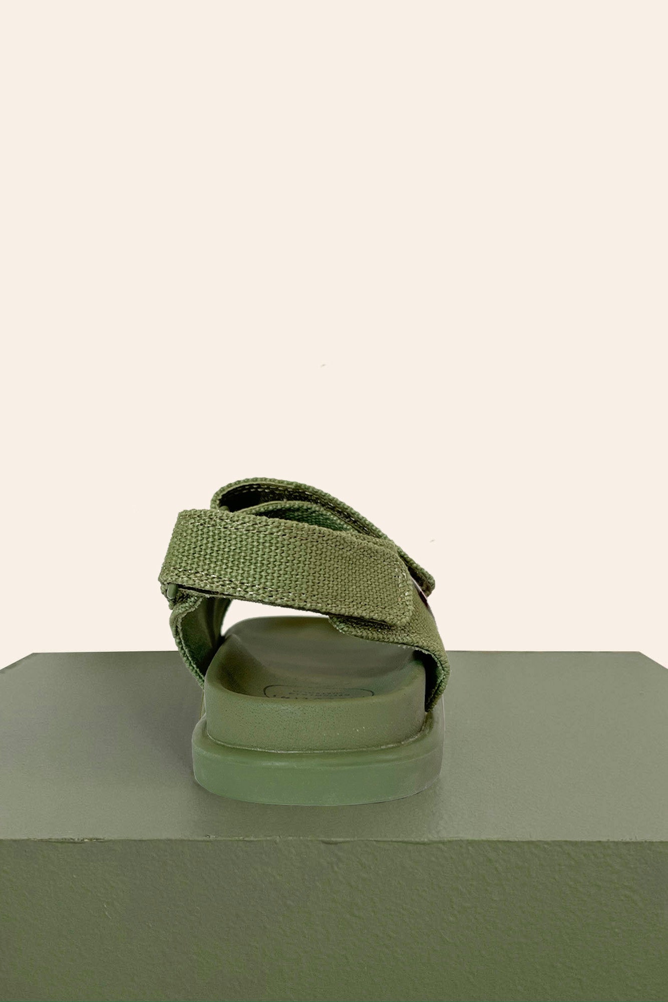 A single khaki sandal from PICCOLINI's PRE-ORDER Original Sandals collection features a textured strap and is crafted from organic cotton, positioned on a matching platform against a cream background. The sandal faces away from the viewer, showcasing its back and sole, emphasizing its podiatrist-recommended comfort.